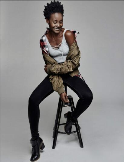 A woman with natural hair, dressed casually in a plaid shirt, white top, and black jeans, smiling while seated on a stool against a plain gray background.